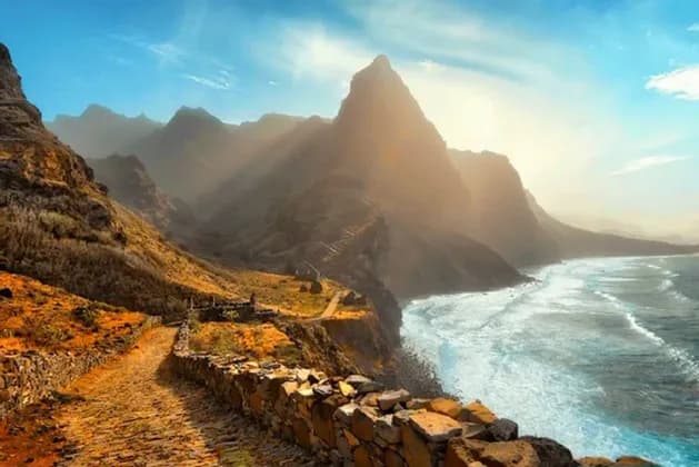 Ein Kopfsteinpflasterweg schlängelt sich entlang einer zerklüfteten Küste neben dem Meer, mit steilen, sonnenbeschienenen Bergen im Hintergrund.