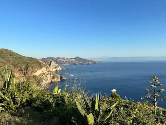 Una vista da una verdeggiante scogliera su una costa frastagliata, un mare blu con barche e isole lontane sotto un cielo sereno.
