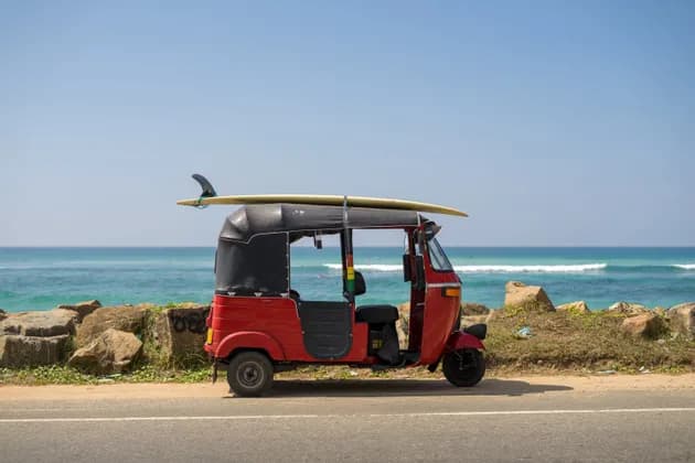 Un tuk-tuk rosso con una tavola da surf legata al tetto è parcheggiato a bordo strada vicino all'oceano sotto un cielo azzurro e limpido.