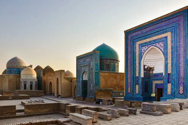 Ein Komplex historischer Mausoleen mit kunstvollen blauen und türkisen Kacheln und Kuppeln in einem steinernen Innenhof bei Sonnenuntergang.