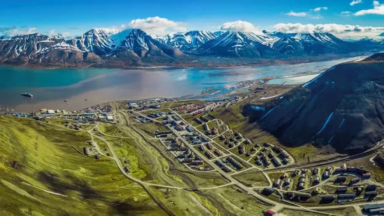 Una vista aérea de un pueblo costero en un valle verde junto a un fiordo, con un telón de fondo de montañas cubiertas de nieve bajo un cielo azul.