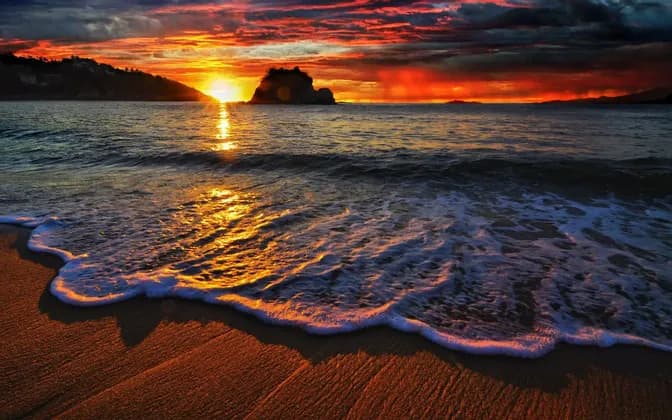 Un'onda si infrange su una spiaggia sabbiosa mentre il sole tramonta sull'oceano, proiettando un bagliore dorato sotto un cielo drammatico e nuvoloso.