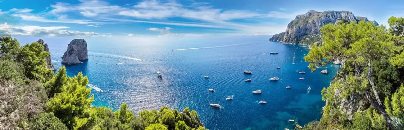Una vista panoramica di una costa rocciosa con un grande faraglione, che si affaccia su un mare azzurro brillante punteggiato da numerose barche e yacht in una giornata di sole.