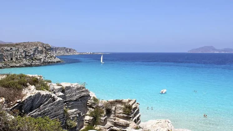 Una vista dall'alto di una costa rocciosa che costeggia una baia turchese con una barca a vela e nuotatori sotto un cielo azzurro e limpido.