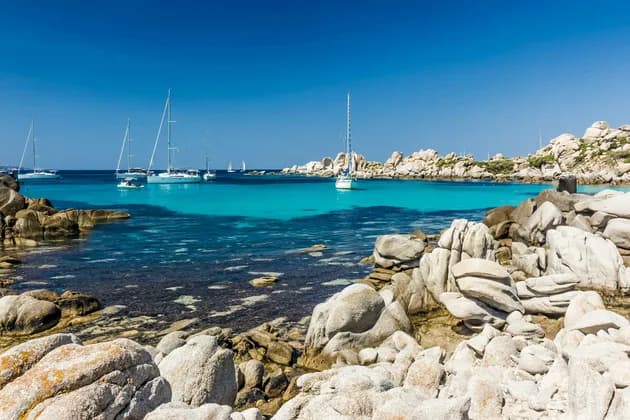 Un littoral rocheux surplombe une baie calme avec des voiliers sur une eau turquoise claire sous un ciel bleu éclatant.