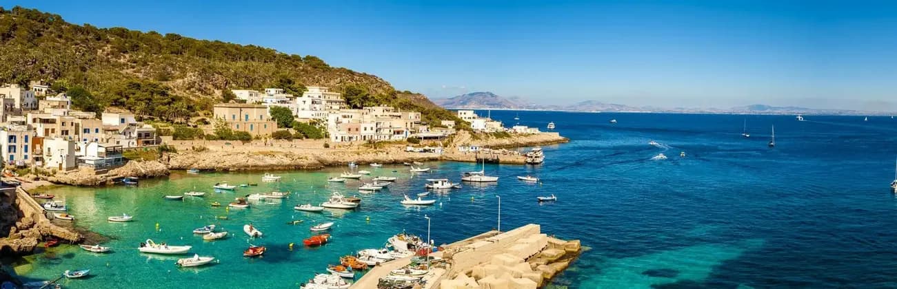 Un villaggio costiero su una verde collina si affaccia su una baia piena di piccole barche su acque turchesi e blu sotto un cielo sereno.
