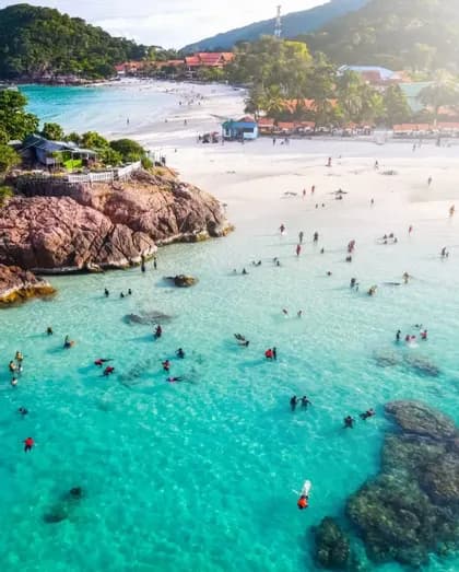 Eine Luftaufnahme von Menschen, die in klarem türkisfarbenem Wasser schwimmen und schnorcheln, neben einem weißen Sandstrand, der von üppig grünen Hügeln gesäumt wird.