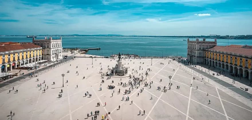 Una vista aérea muestra a personas caminando en una gran plaza pavimentada junto al mar con un monumento central, flanqueada por edificios porticados bajo un cielo azul.