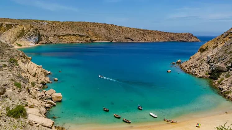 Una vista aerea di un motoscafo che attraversa una baia turchese racchiusa da scogliere rocciose, con diverse piccole imbarcazioni vicino a una spiaggia sabbiosa.
