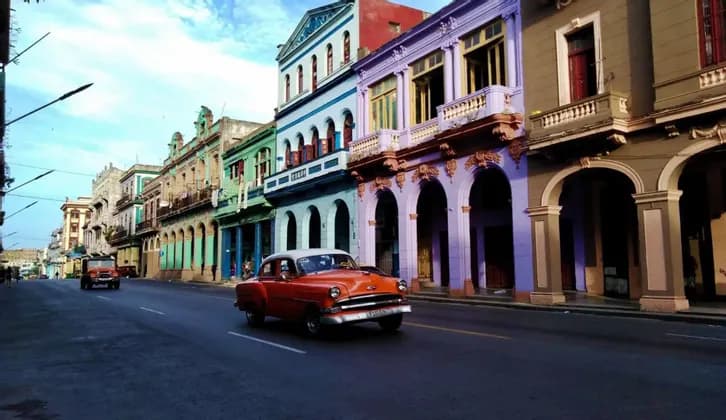 Un'auto d'epoca arancione percorre una strada accanto a colorati edifici coloniali con portici ad arco sotto un cielo blu.