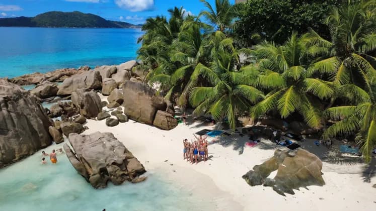 Veduta aerea di un gruppo WeRoad in posa su una spiaggia di sabbia bianca, circondata da grandi massi, palme e acqua turchese.