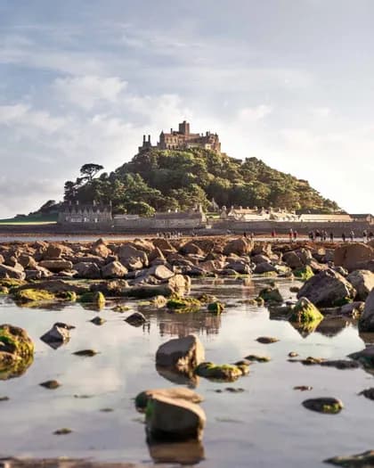 Un castello si erge su un'isola alberata, osservato da una costa rocciosa con piscine naturali in primo piano, di giorno.