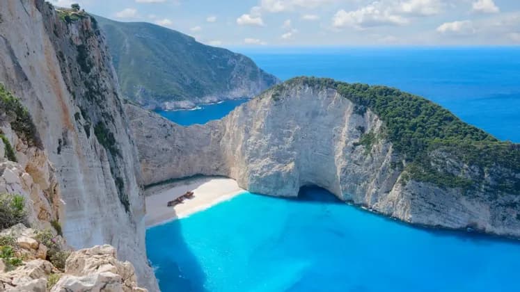 Un relitto arrugginito giace su una spiaggia di sabbia bianca isolata, circondato da imponenti scogliere e acqua turchese sotto un cielo azzurro.