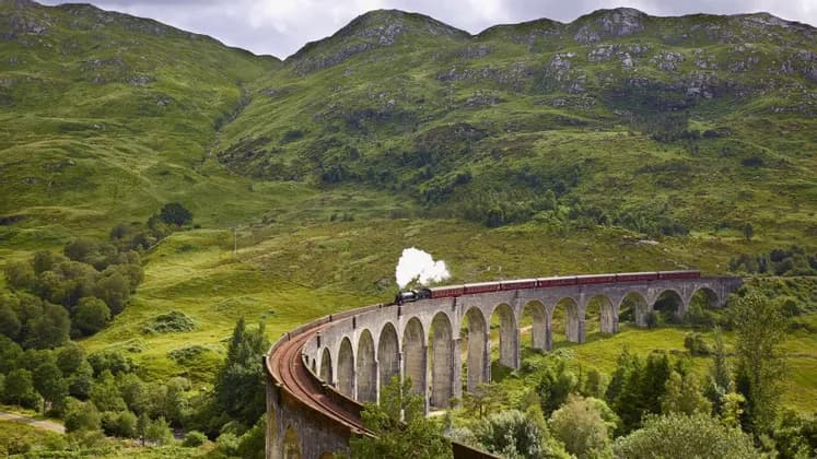 Un treno a vapore con vagoni rossi attraversa un viadotto in pietra curvo in una rigogliosa valle verde circondata da montagne.