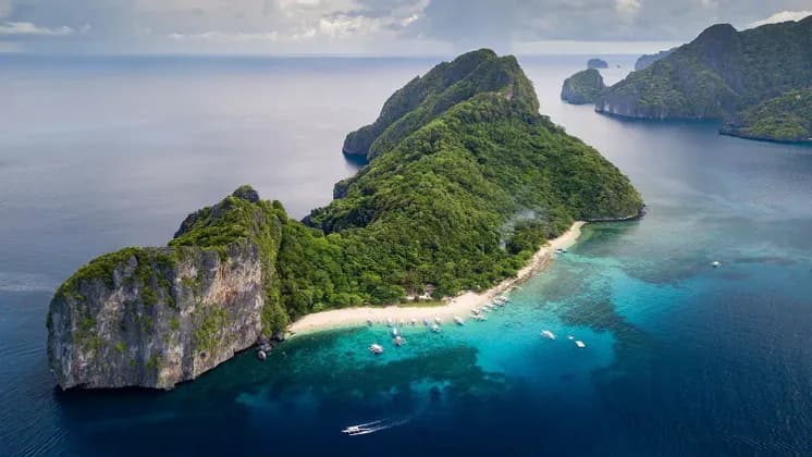 Aerial view of a lush, rocky island featuring a white sand beach with many boats moored in the surrounding turquoise water.