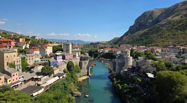 Un vecchio ponte di pietra attraversa un fiume turchese che scorre attraverso una città storica adagiata su una grande collina sotto un cielo azzurro.