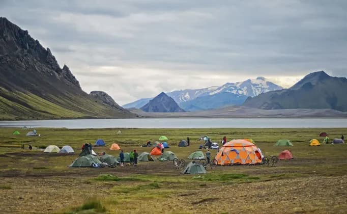 Un voyage de groupe WeRoad avec des tentes colorées et des vélos, campant au bord d'un lac au pied de montagnes escarpées et enneigées.