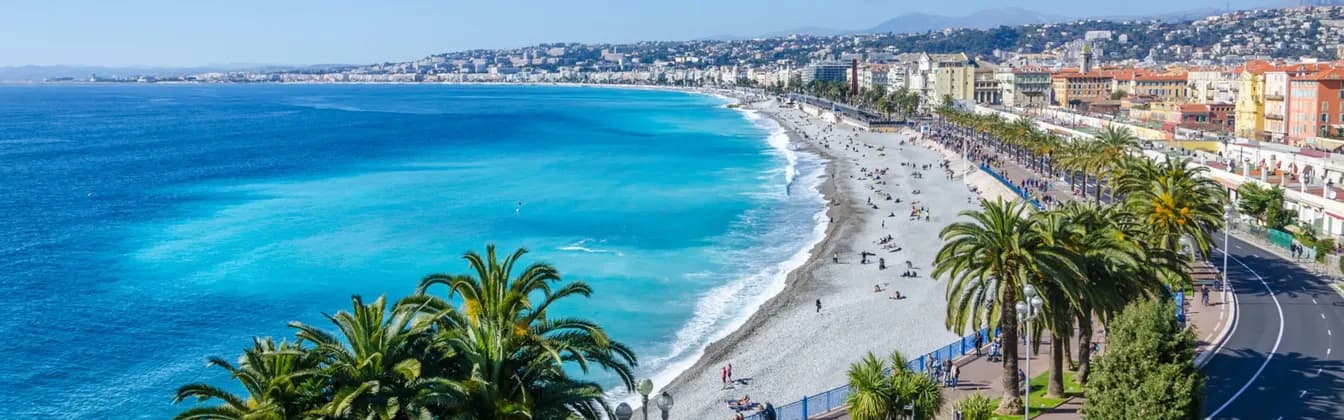 A panoramic view of a coastal city with a long pebble beach, turquoise water, and a promenade lined with palm trees.