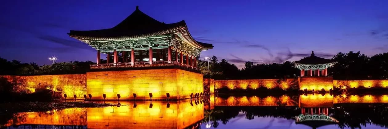 Una tradizionale pagoda asiatica e un muro di pietra illuminati al tramonto, con le loro luci calde che si riflettono nell'acqua immobile.