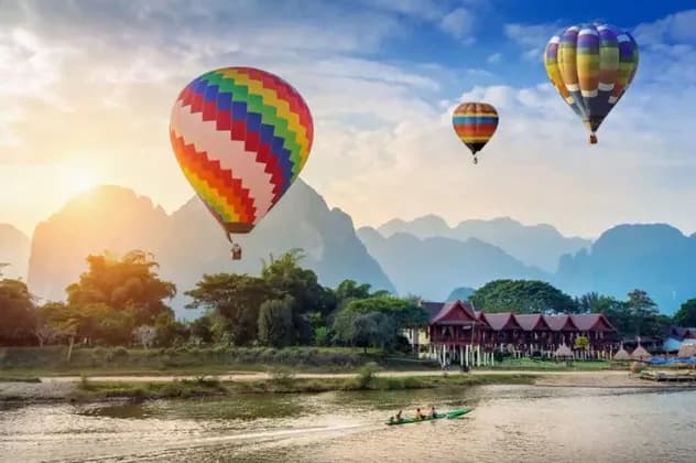 Tre mongolfiere colorate volano su un fiume e montagne lussureggianti all'alba, mentre una piccola barca naviga sull'acqua.