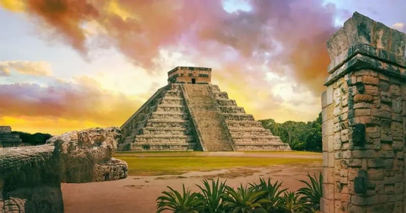 Una piramide di pietra Maya si erge su un campo erboso, incorniciata da antiche rovine di pietra sotto un cielo nuvoloso e colorato al tramonto.