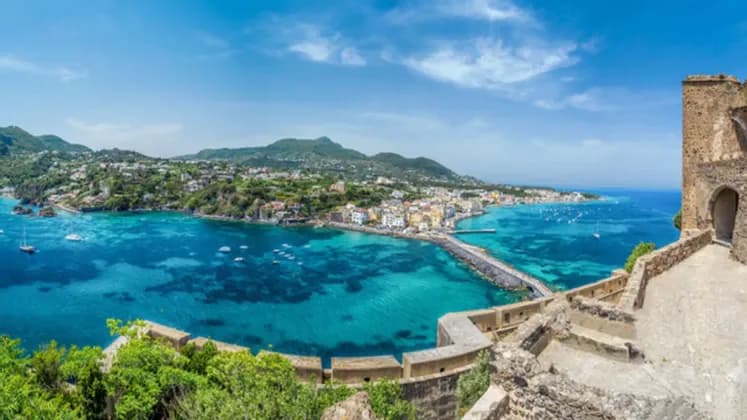 A high-angle, panoramic view of a coastal town and causeway surrounded by turquoise sea, seen from a stone fortification.