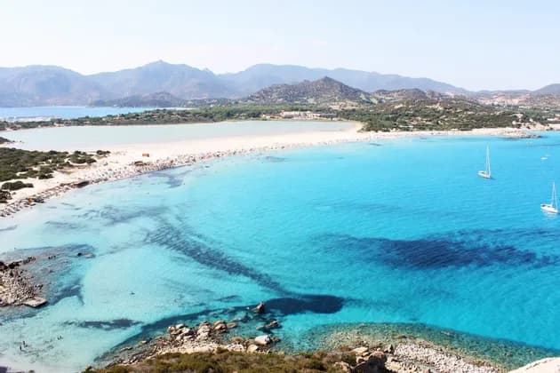 An aerial view of a crowded white sand beach bordering vibrant turquoise water with sailboats, with mountains in the background.
