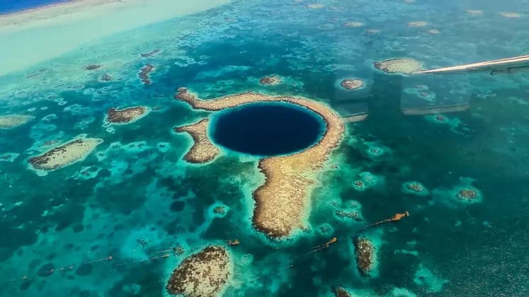 An aerial view from an airplane of a large, dark blue marine sinkhole surrounded by a coral reef in a shallow turquoise ocean.