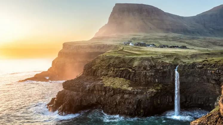 Una cascata si getta da una verde scogliera erbosa nell'oceano, con un villaggio sullo sfondo al tramonto.
