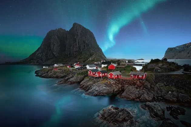 Case rosse su una costa rocciosa si ergono sotto un'imponente montagna mentre l'aurora boreale risplende nel cielo stellato notturno.