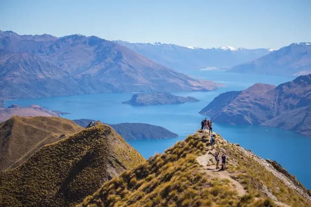 A WeRoad group trip stands on a grassy mountain peak, overlooking a large blue lake surrounded by mountain ranges.