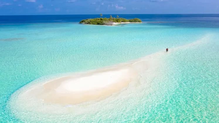Una vista aerea di due persone che camminano su un banco di sabbia bianca che si estende attraverso acque cristalline color turchese verso una piccola isola tropicale.