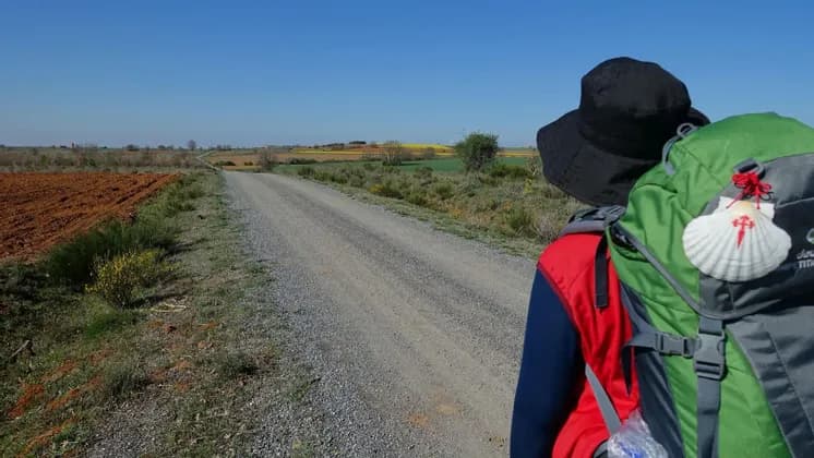 Una persona con uno zaino e una conchiglia del Cammino di Santiago cammina su una strada sterrata attraverso un paesaggio rurale.
