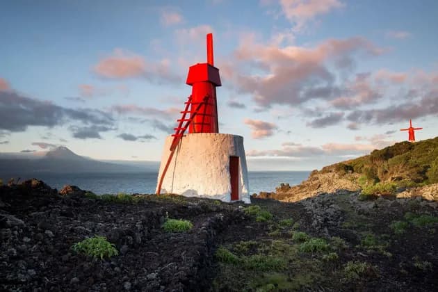Un faro bianco e rosso su una costa vulcanica, con una montagna lontana e un mulino a vento rosso sullo sfondo.