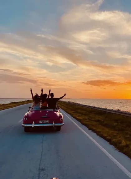 Un viaggio di gruppo WeRoad visto da dietro, alla guida di una decappottabile vintage rosa su una strada costiera durante un tramonto dorato.
