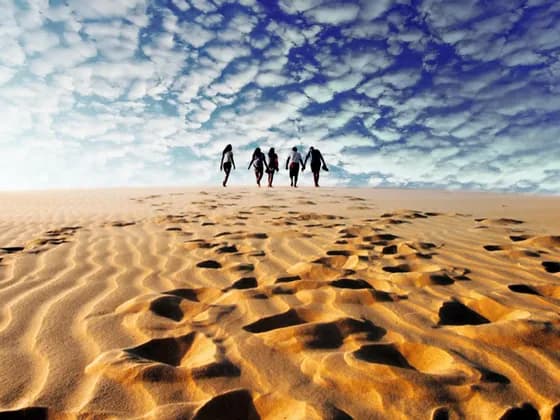 Un voyage de groupe WeRoad traverse une vaste dune de sable, laissant des empreintes dans le sable sous un ciel bleu nuageux.