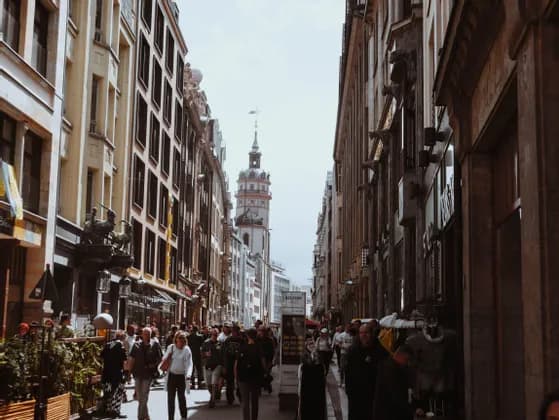 Eine Menschenmenge geht eine enge Stadtstraße entlang, die von hohen, historischen Gebäuden und einem fernen Glockenturm gesäumt ist.