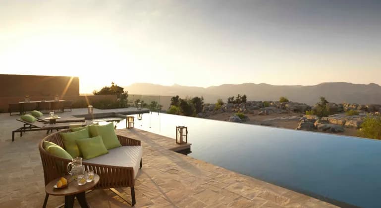 A modern patio with an infinity pool and lounge furniture overlooking a rocky mountain landscape at sunset.