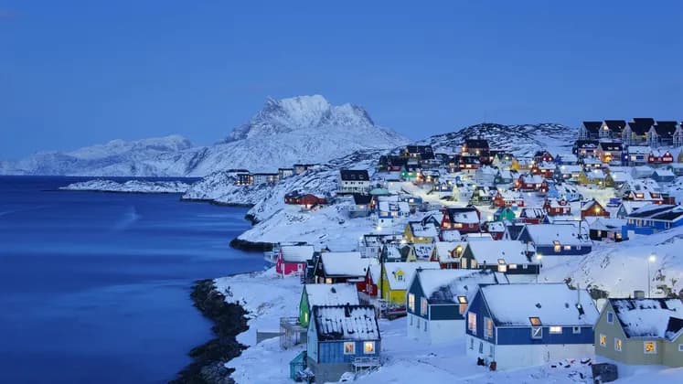 Un villaggio costiero innevato di case colorate con luci accese al tramonto, incastonato tra le montagne e un mare calmo.