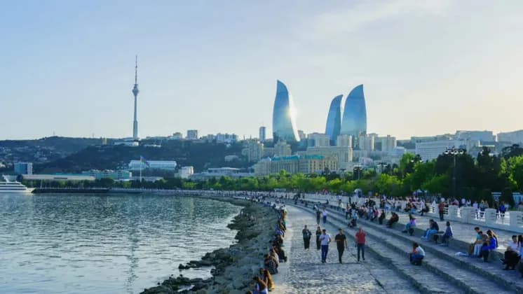 Eine Menschenmenge entspannt sich auf einer breiten, sonnigen Uferpromenade, im Hintergrund eine moderne Stadtsilhouette mit geschwungenen Glastürmen.