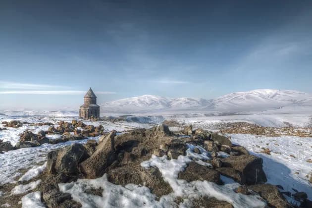 Une église en pierre au toit conique se dresse dans un vaste paysage enneigé, avec des ruines rocheuses et des montagnes en arrière-plan.