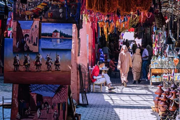 People walk through a bustling market alley filled with paintings for sale and colorful dyed yarns hanging overhead.