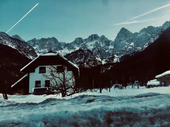 Una casa bianca con il tetto innevato adagiata in una valle innevata ai piedi di una catena montuosa frastagliata.