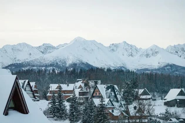 Un village de chalets et de maisons enneigés au pied d'une forêt de pins, avec une grande chaîne de montagnes enneigées en arrière-plan.