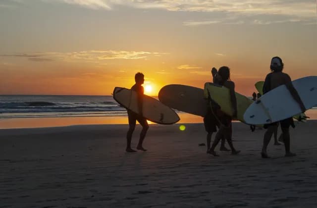 Un gruppo WeRoad di surfisti in controluce al tramonto, che camminano sulla spiaggia con le loro tavole da surf.