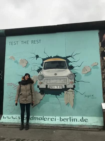 Una donna con un cappotto invernale sorride mentre posa davanti a un grande murale di un'auto Trabant che sfonda un muro.