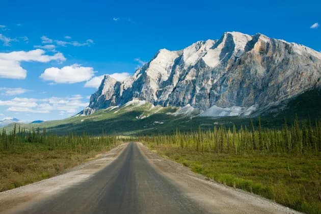 Una strada sterrata diritta attraversa una valle verde con alberi verso una grande catena montuosa rocciosa sotto un cielo azzurro con nuvole.