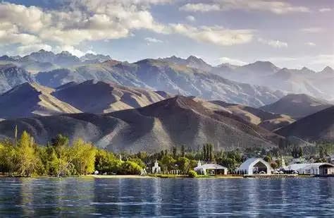 Un lago en calma se extiende frente a una orilla con árboles verdes y edificios blancos, con una vasta y ondulante cordillera de fondo bajo un cielo parcialmente nublado.
