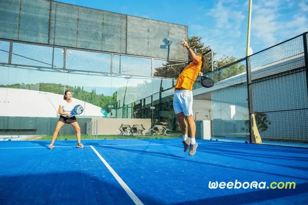 Un uomo con una maglietta arancione salta per servire mentre gioca a padel con una donna su un campo blu brillante all'aperto, sotto un cielo sereno.