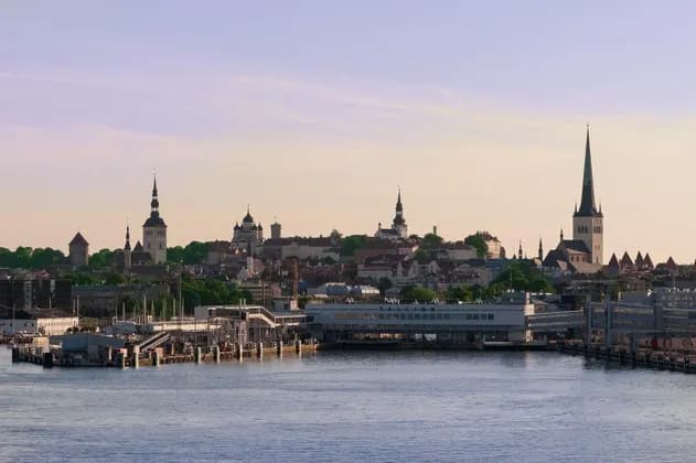 Lo skyline di una storica città europea, con guglie e torri di chiese, si erge dietro un moderno porto in riva all'acqua.